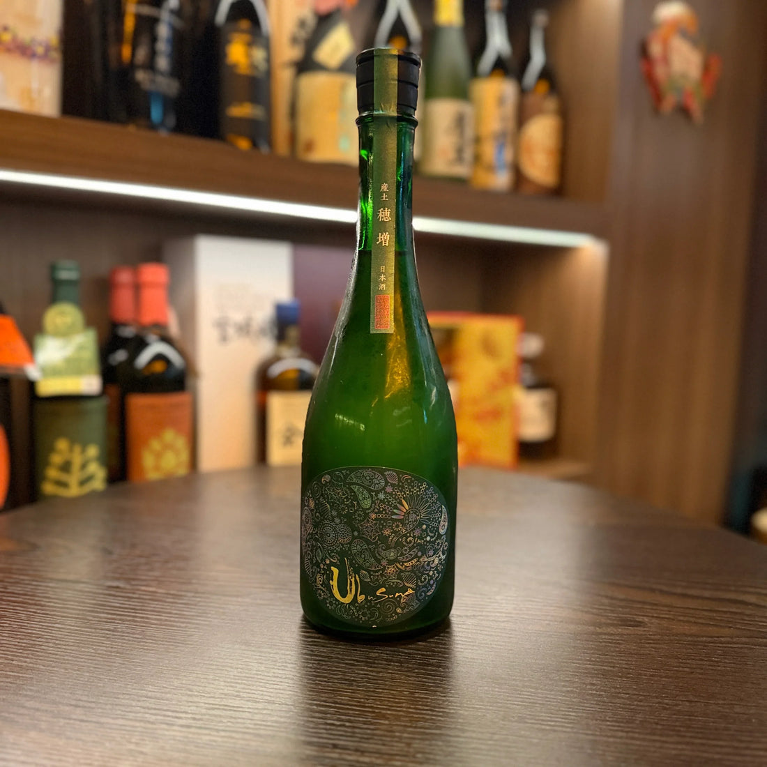 一瓶綠色玻璃瓶裝的日本清酒，瓶身帶有精美圖案與日文標籤，擺放在深色木桌上。